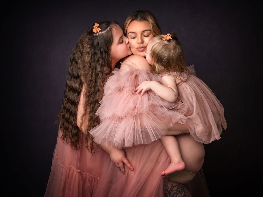 Canterbury Photographer. pregnant lady wearing a pink dress being kissed by her two daughters in matching dresses.